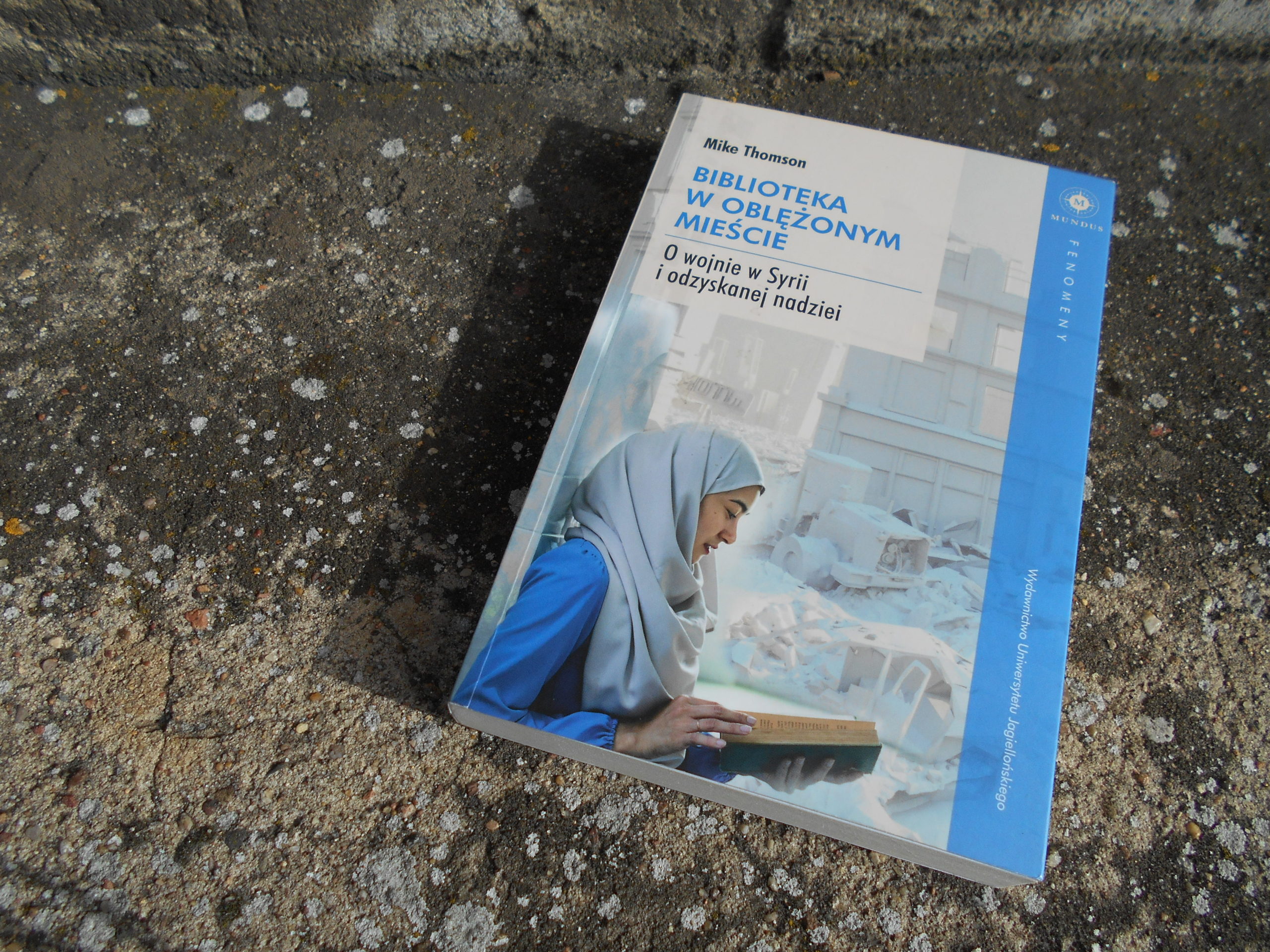 zdjęcie przedstawiające szaro-niebieską książkę "Biblioteka w oblężonym mieście. O wojnie w Syrii i odzyskanej nadziei", leżącą na chodniku